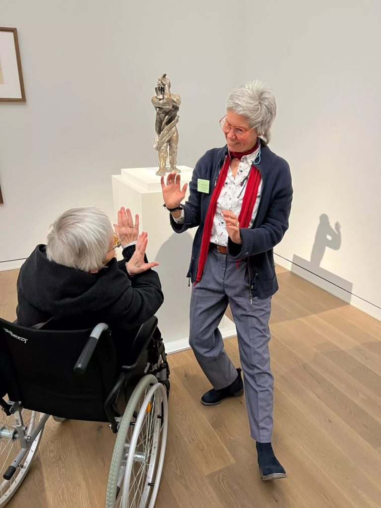 Kunstvermittlerin stellt die Geste einer Skulptur zusammen mit einer Person im Rollstuhl nach.